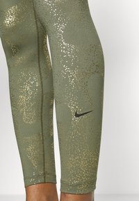 Leggings verde oliva con motivo a pois dorati, caratterizzati da un logo Nike nero. Texture liscia e design aderente che si restringe alla caviglia.