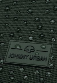 Tmavozelená textúrovaná látka s kvapkami vody, s tmavým štítkom, na ktorom sú tučnými písmenami napísané "JOHNNY URBAN".