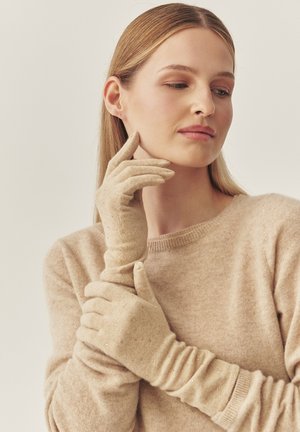 Bēšas adītas cimdi no mīksta auduma ar teksturētu virsmu, rotāti ar mazām dekoratīvām pērlītēm. Dizainā ir cieši pieguļošs aproce.