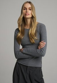 Szara koszulka z długim rękawem o dopasowanym kroju, okrągłym dekolcie i miękkiej fakturze. W połączeniu z ciemnymi, szerokimi spodniami.