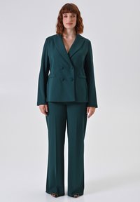 Donkergroene op maat gemaakte blazer met een dubbelgeknoopte sluiting, gecombineerd met bijpassende wijde pantalons van een gladde stof.