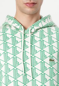 Felpa con cappuccio caratterizzata da un tessuto bianco testurizzato con un motivo geometrico verde, accentuata da un piccolo logo di un coccodrillo verde sul petto.