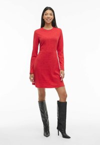 Robe longue à manches longues rouge avec motif texturé, ourlet évasé et coupe slim. Associée à des bottes en cuir noires montant jusqu'aux genoux avec des bouts pointus.