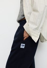 Ręka w kieszeni, ubrana w granatowe spodnie dresowe z logo P08 Studio Rotterdam, w połączeniu z szarym swetrem i białą koszulą w paski na guziki.