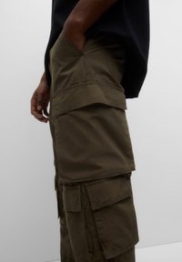 Pantalon cargo vert avec plusieurs poches, fermeture par bouton et coupe ample. Le tissu est lisse avec des détails de couture visibles.