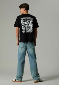 Svart t-shirt med vitt grafiskt text på baksidan, som visar evenemangsdetaljer. Kombinerad med ljusblå baggy jeans och vita sneakers.