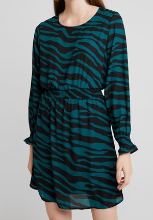 Femme portant une robe à manches longues bleu canard et noire avec des rayures zébrées, une taille élastique et des poignets à volants, coupée mi-cuisse.