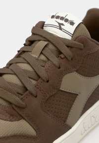 Brown athletic shoe featuring a mix of suede and mesh materials, textured accents, and rounded toe design with a logo on the tongue.