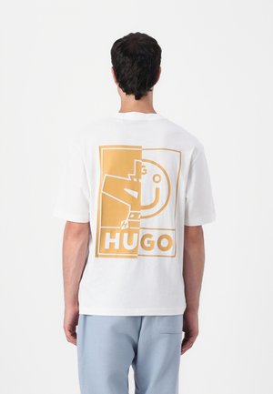 Mężczyzna ubrany w białą koszulkę z dużą żółto-białą grafiką i napisem "HUGO" z tyłu, w połączeniu ze spodniami w jasnoniebieskim kolorze.