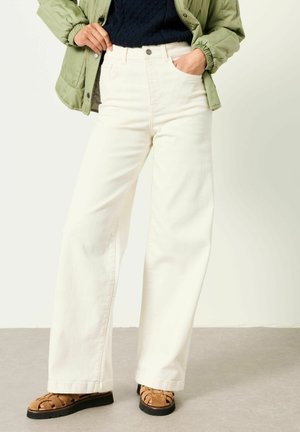 Személy, aki magas derekú, bőszárú fehér nadrágot, fekete pulóvert, zöld zakót és barna fonott szandált visel, szürke padlón áll.