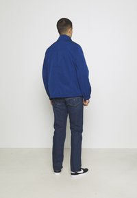 Blå lättviktsjacka med ståkrage, elastiska muddar och avslappnad passform, kombinerad med mörka denimjeans och svarta sneakers.