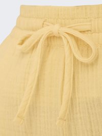 Gule teksturerede stofbukser med elastisk talje og bundet snøre foran, der viser en del af en sidelomme.
