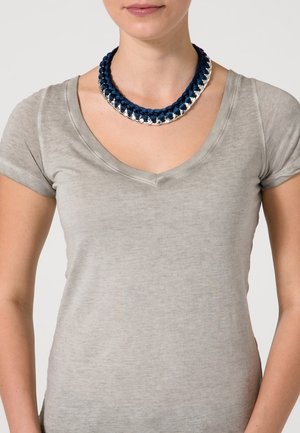 Kobieta nosząca pleciony naszyjnik łączący granatową tkaninę z srebrnymi metalowymi ogniwami, na jasnoszarej koszulce z dekoltem w serek.