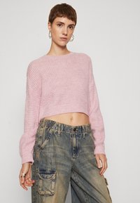 Ljusrosa, kort stickad tröja med ett strukturerat mönster; matchad med lösa, blekta blå jeans med dragkedja i midjan och fickor.