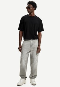 Muškarac s kovrčavom kosom nosi sunčane naočale, crnu preveliku majicu, svijetlosive cargo traperice i bijele tenisice, stojeći na bijeloj pozadini.