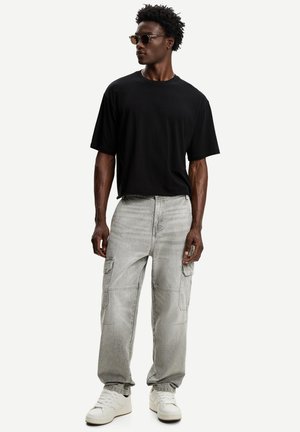 Mężczyzna z kręconymi włosami, noszący okulary przeciwsłoneczne, czarny oversize'owy t-shirt, jasnoszare spodnie cargo i białe sneakersy, stojący na białym tle.