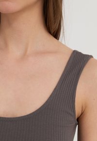 Grå ribbet tank topp med en rundet utringning, med brede skulderstropper og en tettsittende silhuett. Glatt tekstur på hudfarget bakgrunn.