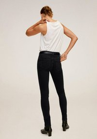 Nainen, joka on pukeutunut valkoiseen hihattomaan pueeseen, mustiin skinny-farkkuihin, joissa on vyö, ja mustiin nilkkabootseihin, seisoo yksinkertaista taustaa vasten selkä kameraan päin.