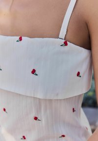 Weißes ärmelloses Oberteil mit bestickten roten Blumen und grünen Stängeln, das ein geschichtetes Design und eine vertikale Streifenstruktur aufweist.