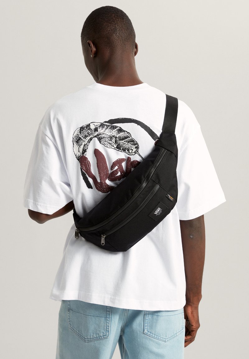 Musta olkalaukku, jossa teksturoitu pinta, kaksi vetoketjullista lokeroa, säädettävä hihna, käytetty rinnan yli valkoisen käärmekuvioisen t-paidan päällä.