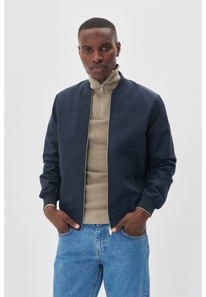 Granatowa kurtka bombowa z zamkiem z przodu, prążkowanymi mankietami i kołnierzem, w połączeniu z beżowym swetrem zapinanym na zamek oraz jasnoniebieskimi dżinsami. Gładka tekstura.