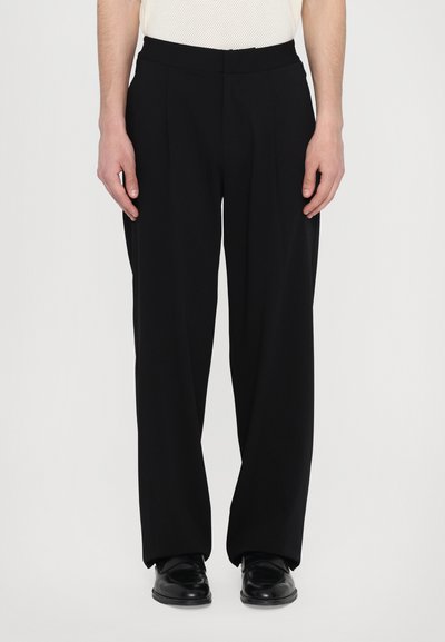Black, wide-leg trousers in a smooth fabric with a flat waistband, featuring front pleats. Paired with shiny black footwear.