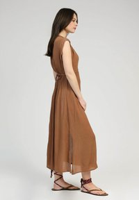 Robe marron, longueur cheville avec manches courtes et fentes latérales. Tissu léger et froissé ; détail de taille noué ; associée à des sandales à lanières.