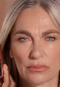 Close-up van een lichtgekleurde persoon met lang, steil, blond haar die een bruine lippenstifttube vasthoudt. De huid vertoont fijne lijntjes en een natuurlijke make-uplook.