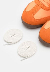 Pomarańczowe zamszowe i siateczkowe sneakersy z zaokrąglonym noskiem, uzupełnione dwoma białymi sznurówkami, na neutralnym tle.