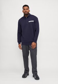 Granatowy sweatshirt zapinany na zamek z wysokim kołnierzem, z białym logo na lewej piersi. W połączeniu z szarymi spodniami cargo i czarnymi sneakersami.