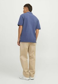 Muškarac stoji okrenut leđima, nosi plavu majicu s kratkim rukavima, bež hlače i bijele tenisice na jednolično svijetloj pozadini.