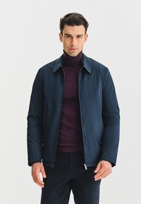 Granatowa, lekka kurtka z klasycznym kołnierzem i zapięciem na zamek, zestawiona z ciemnofioletowym golfem. Gładka faktura, minimalne zdobienia.