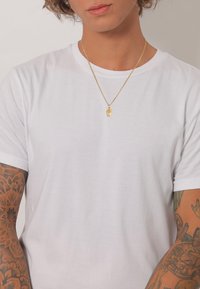 Guld halskæde med et håndformet vedhæng, båret over en ensfarvet hvid t-shirt. Tatoveringer synlige på den ene arm med intrikate blomstermønstre.