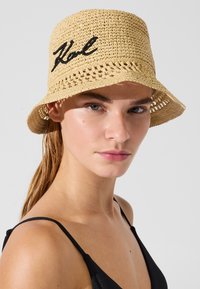 Looduslikust õlgedest kaabu laiade äärega, esiküljel musta tikandiga tekst "Karl". Avatud koega tekstuur, helebeež värv.