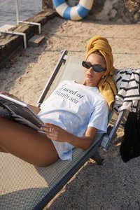 T-shirt en tie-dye bleu clair avec le texte « J'ai besoin de vitamine mer », portée par une personne allongée sur un transat, tenant un magazine, avec une serviette jaune.