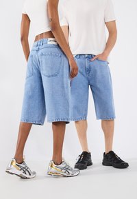 Light blue denim shorts with a relaxed fit, featuring back pockets and a raw hem. Worn with sporty shoes—silver and gold on one, black on the other.