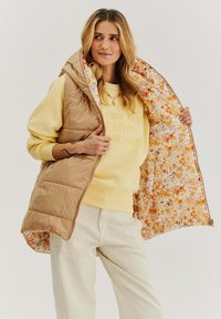 Hnedá puff vest s kvetinovou podšívkou, nosená otvorená cez žltý sveter. Obsahuje zips vpredu a bočné vrecká, skombinovaná so svetlými nohavicami.