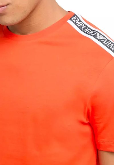 Camiseta de algodón naranja con cuello redondo, que presenta una franja de marca en blanco y negro en el hombro. Textura suave y diseño ajustado.