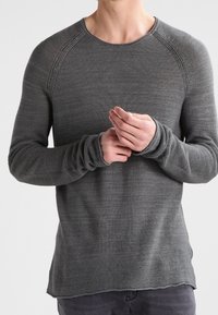Personne portant un pull ajusté à manches longues gris foncé texturé, les mains jointes au niveau de la taille.