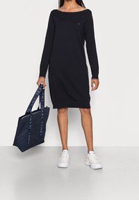 Navy långärmad klänning med avskuren axeldesign, minimal märkning på bröstet. Åtföljd av en mörk väska med logoband och vita sneakers.