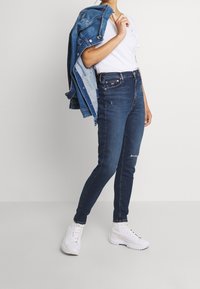 Jeans skinny de mezclilla azul con cintura alta; presenta un detalle rasgado y un ajuste elegante. Combinados con una camiseta blanca y una chaqueta de mezclilla, acompañados de botas blancas.