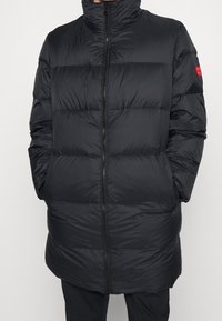 Chaqueta acolchada negra con cuello alto, cierre de cremallera y patrón acolchado horizontal. Cuenta con bolsillos laterales y un acento rojo de marca en la manga.