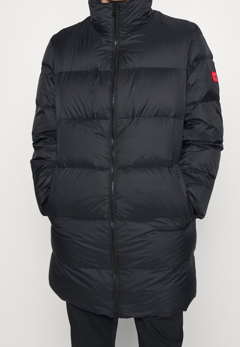 Schwarze Steppjacke mit hohem Kragen, Reißverschluss und horizontalem Steppmuster. Verfügt über seitliche Taschen und einen markanten roten Akzent am Ärmel.