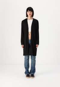 Black long cardigan made of soft fabric, open front, with long sleeves. Pair of blue jeans and black sneakers with beige soles.