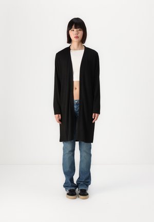 Cardigan long noir en tissu doux, à ouverture frontale, avec des manches longues. Un pantalon en jean bleu et des baskets noires avec des semelles beiges.