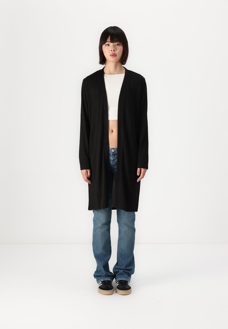 Black long cardigan made of soft fabric, open front, with long sleeves. Pair of blue jeans and black sneakers with beige soles.