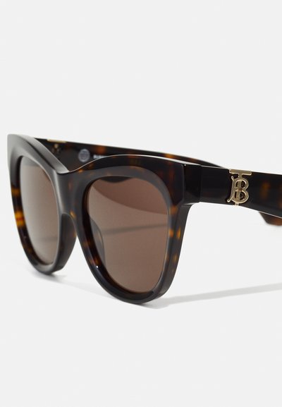 Lunettes de soleil écaille de tortue marron avec une monture large et angulaire. Présente des verres sombres et un accent logo en ton doré sur la branche. Texture lisse.