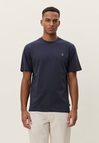 Tamsiai mėlynas medvilninis marškinėlis su apvalia iškirpte, trumpomis rankovėmis ir mažomis baltomis grafikomis krūtinės srityje. Lygus paviršius.