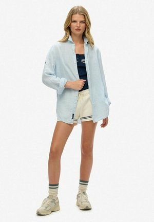 Camicia azzurro chiaro a maniche lunghe sopra una canotta scura, abbinata a pantaloni corti crema. Completata con sneakers beige e calze a righe.