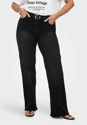 Donna che indossa jeans neri a vita alta a gamba larga con cintura, t-shirt bianca con grafica e sandali neri open-toe, mani nelle tasche.